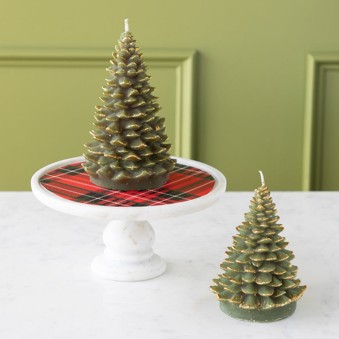 Two tree shaped candles on a table––one on a pedestal with a red plaid pattern underneath and the other on the table.