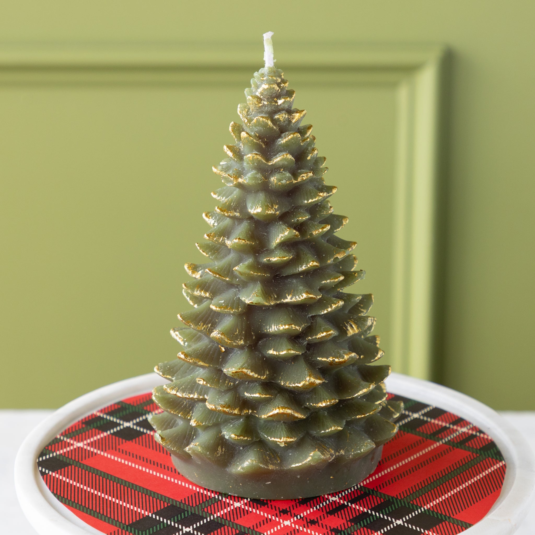 Green candle shaped like a Christmas tree with gold tips on a pedestal with a red plaid pattern underneath.