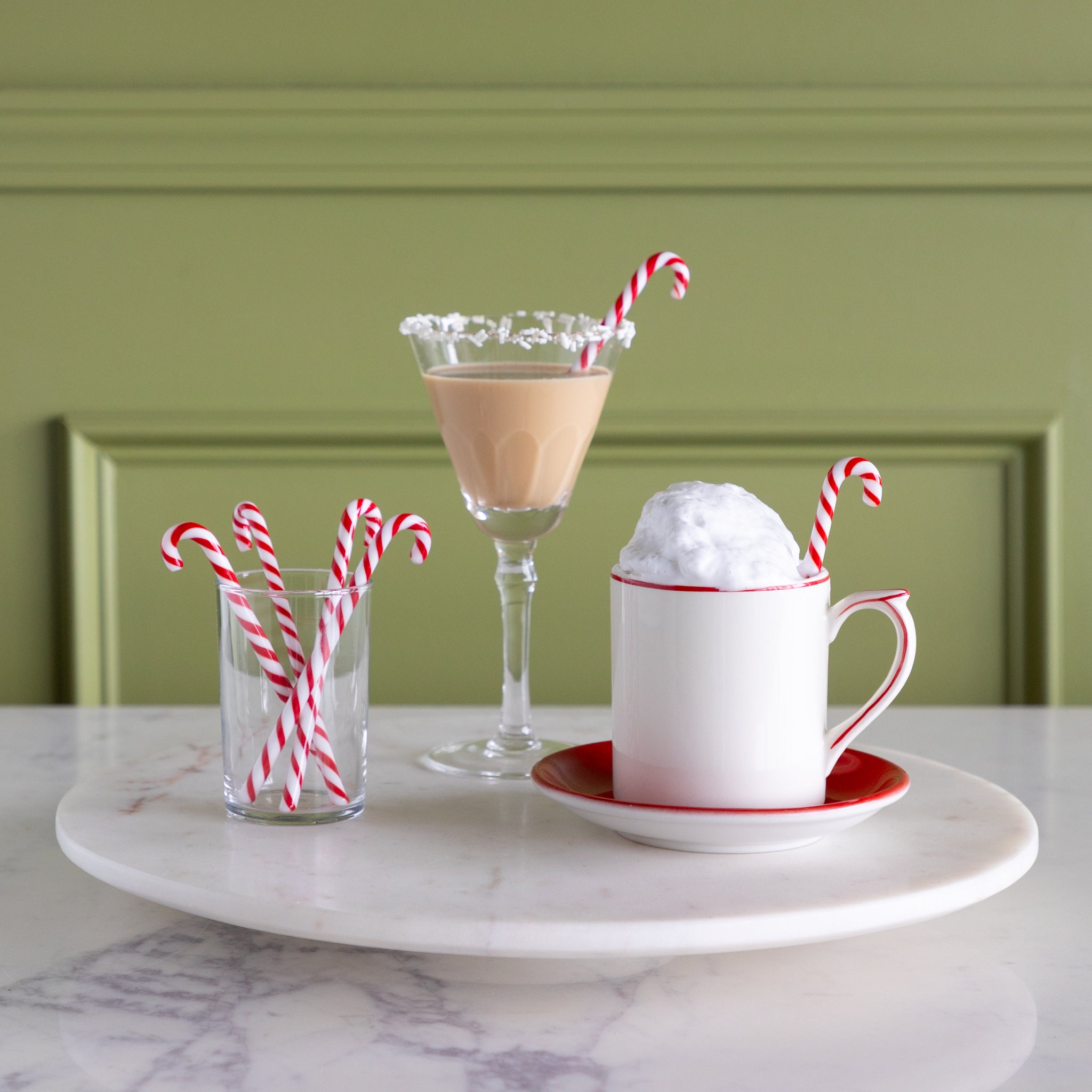 A mug with whipped cream, a mocha cocktail and a small glass with candy cane stir sticks in them on a marble lazy susan.