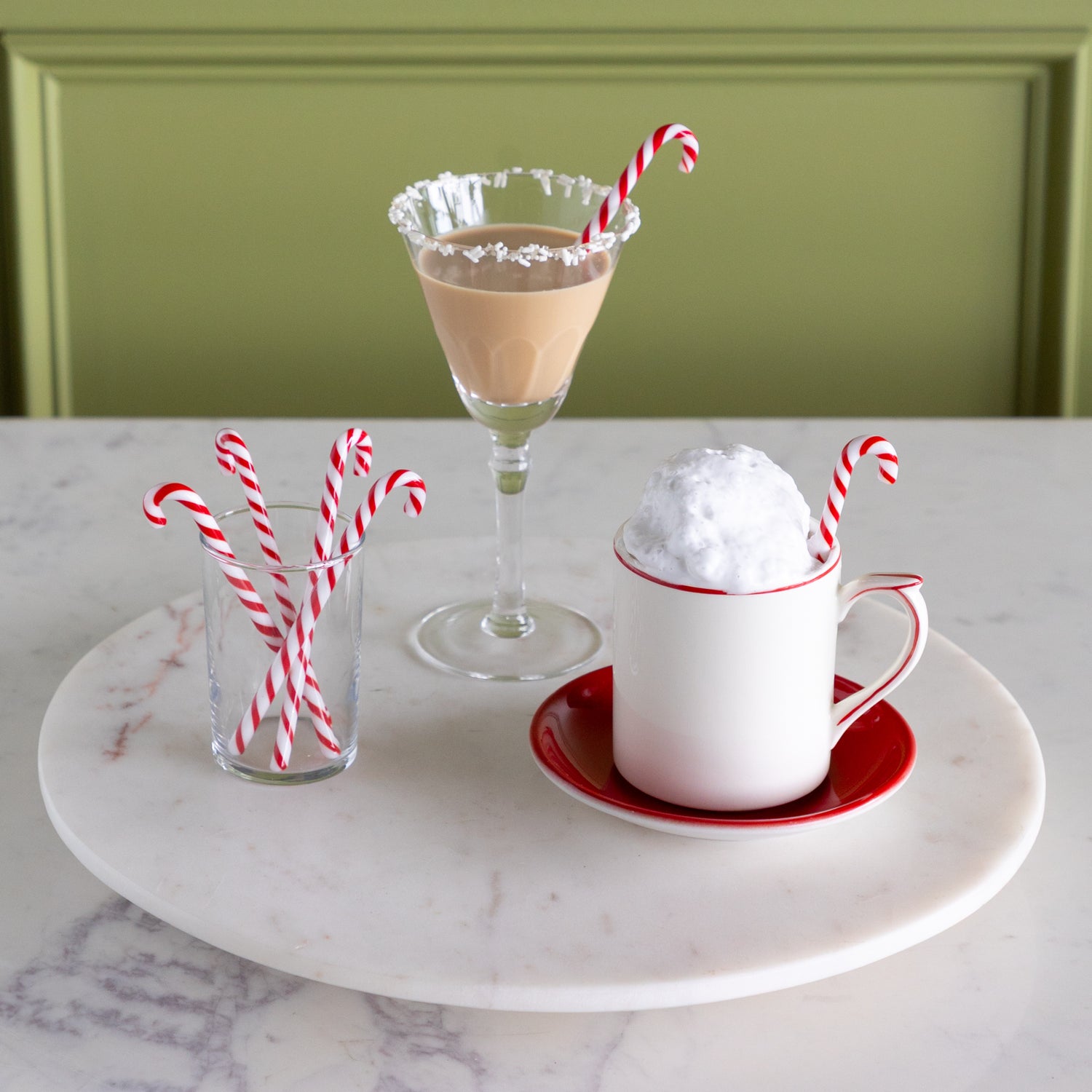 A mug with whipped cream, a mocha cocktail and a small glass with candy cane stir sticks in them on a marble lazy susan.