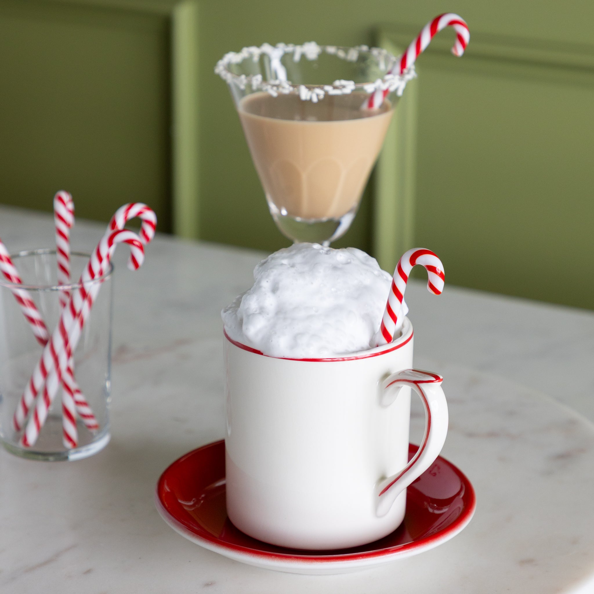 A mug with whipped cream, a mocha cocktail and a small glass with candy cane stir sticks in them on a marble lazy susan.