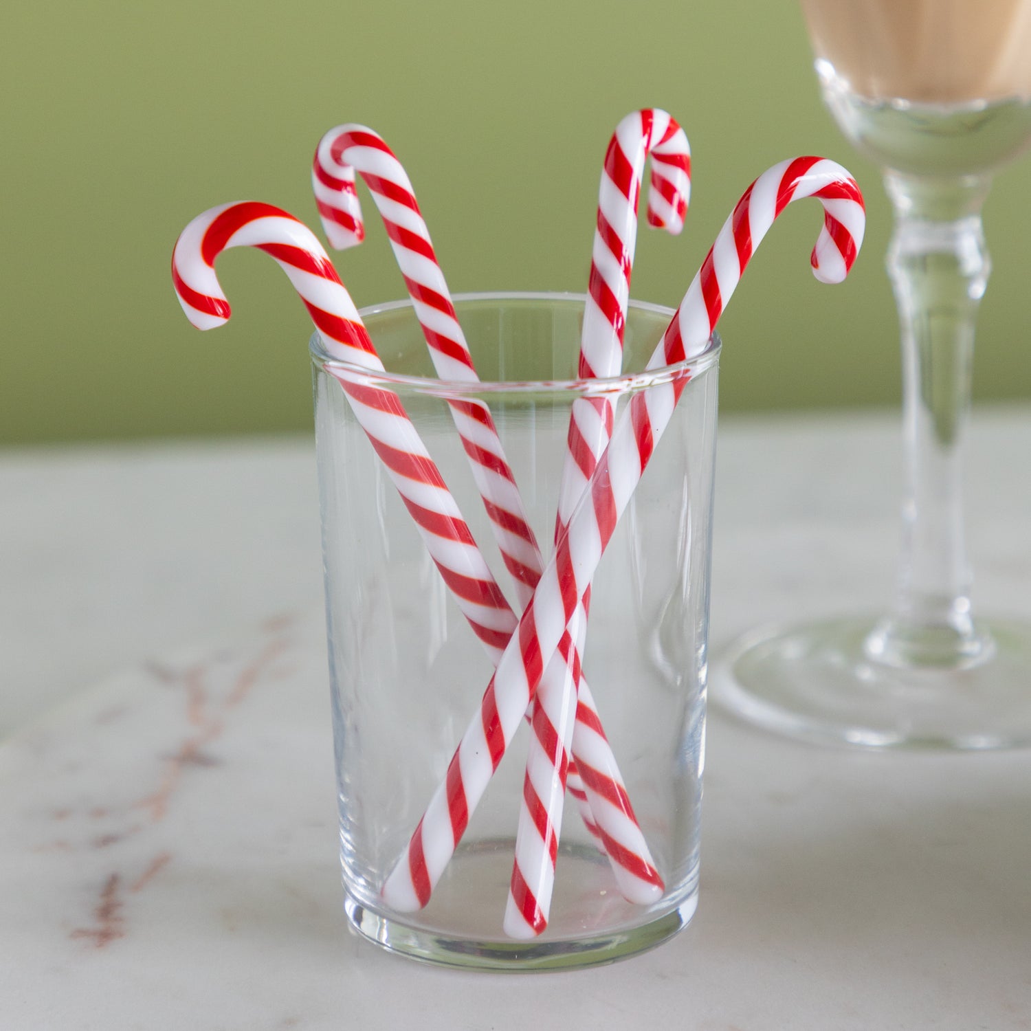 Red and white candy cane stir sticks in a clear glass on a marble surface with a green background.