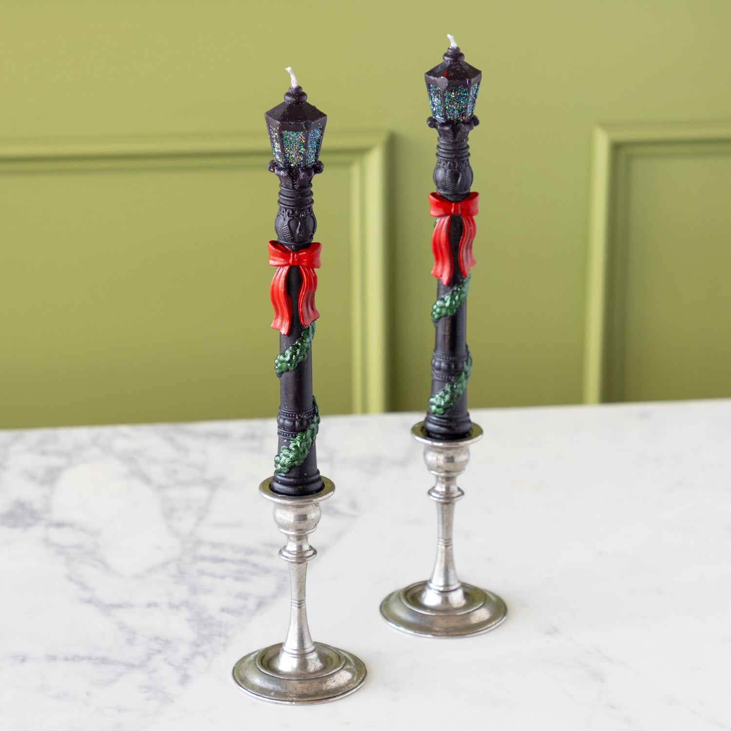 Two Lamppost Taper Candles with a green wreath wrapped around the post, red bows and glittery panes in silver candle holders.