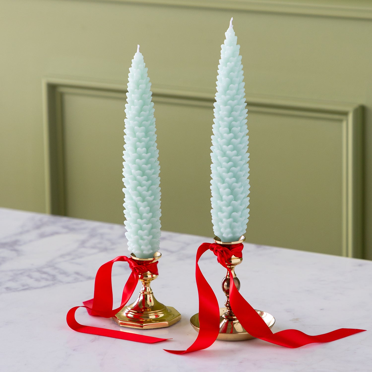 Two Mint Large Pinecone Taper Candles in gold candlesticks with red ribbons on a marble surface.