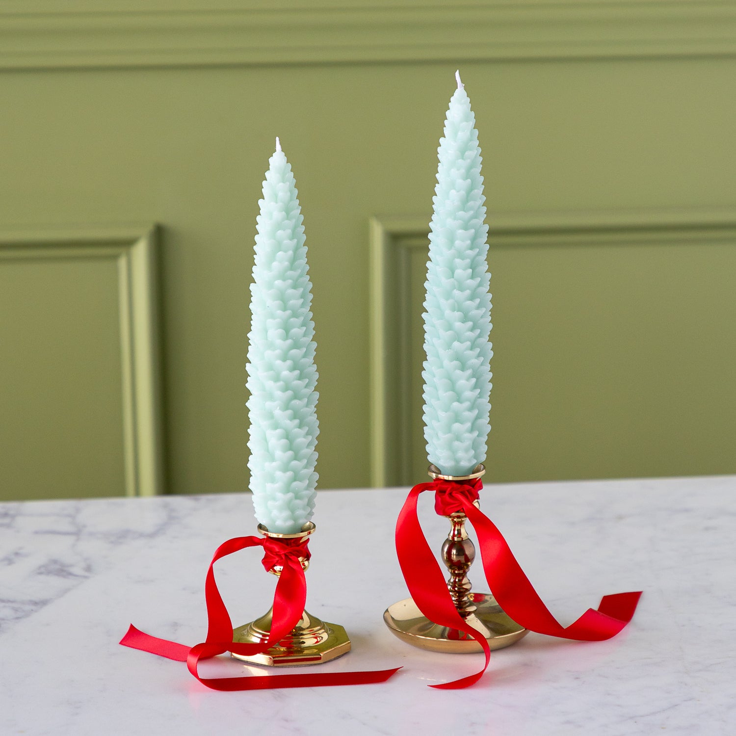 Two Mint Large Pinecone Taper Candles in gold candlesticks with red ribbons on a marble surface.