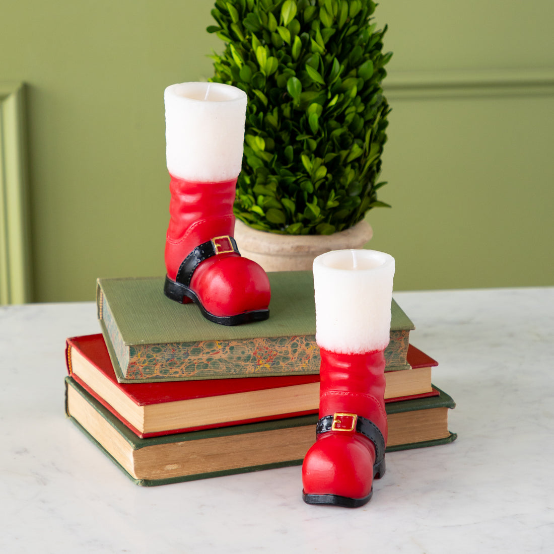 Two Santa Boot Shaped Candles––one on a stack of books and one next to it with a topiary plant in the background.