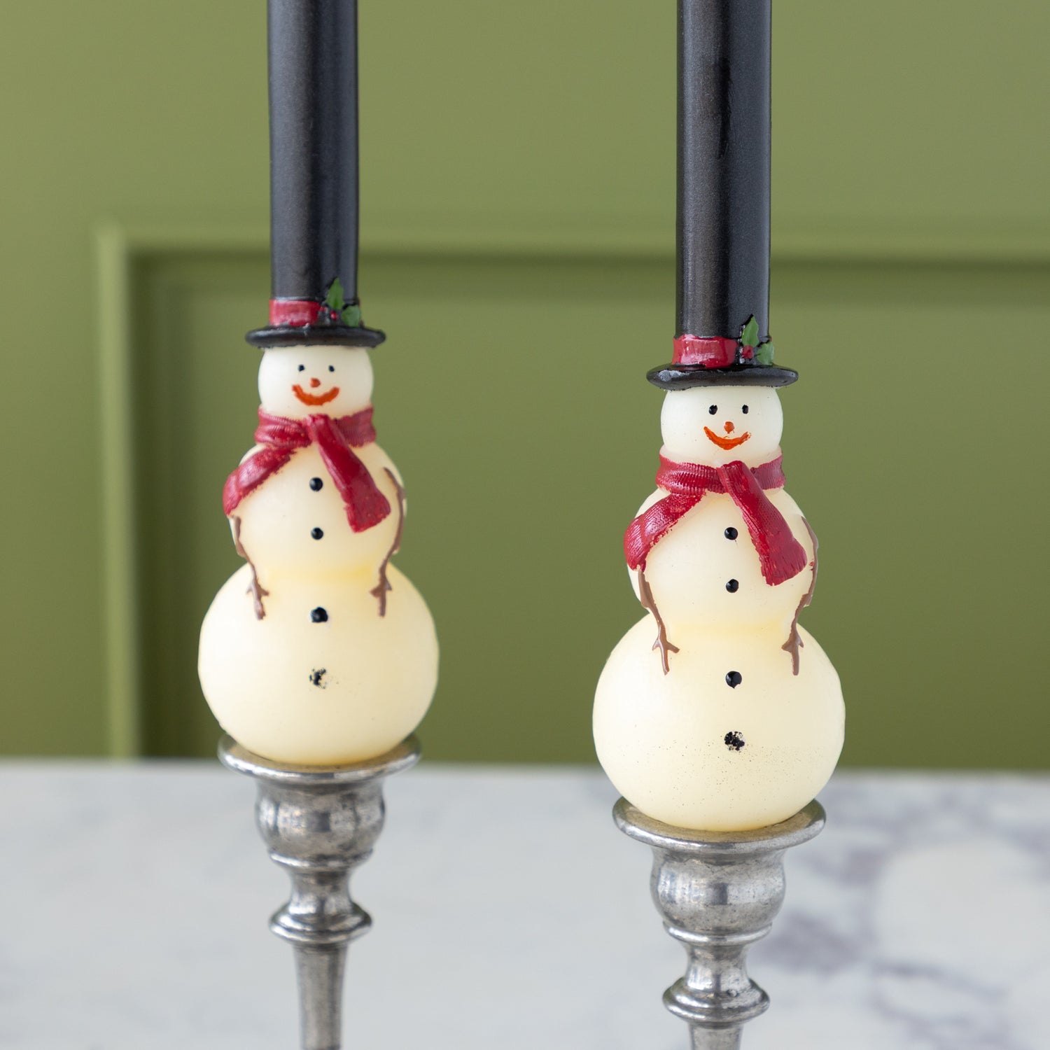 Two Snowman Taper Candles with tall black hats adorned with a red band and holly, red scarves, black buttons and smiley faces.