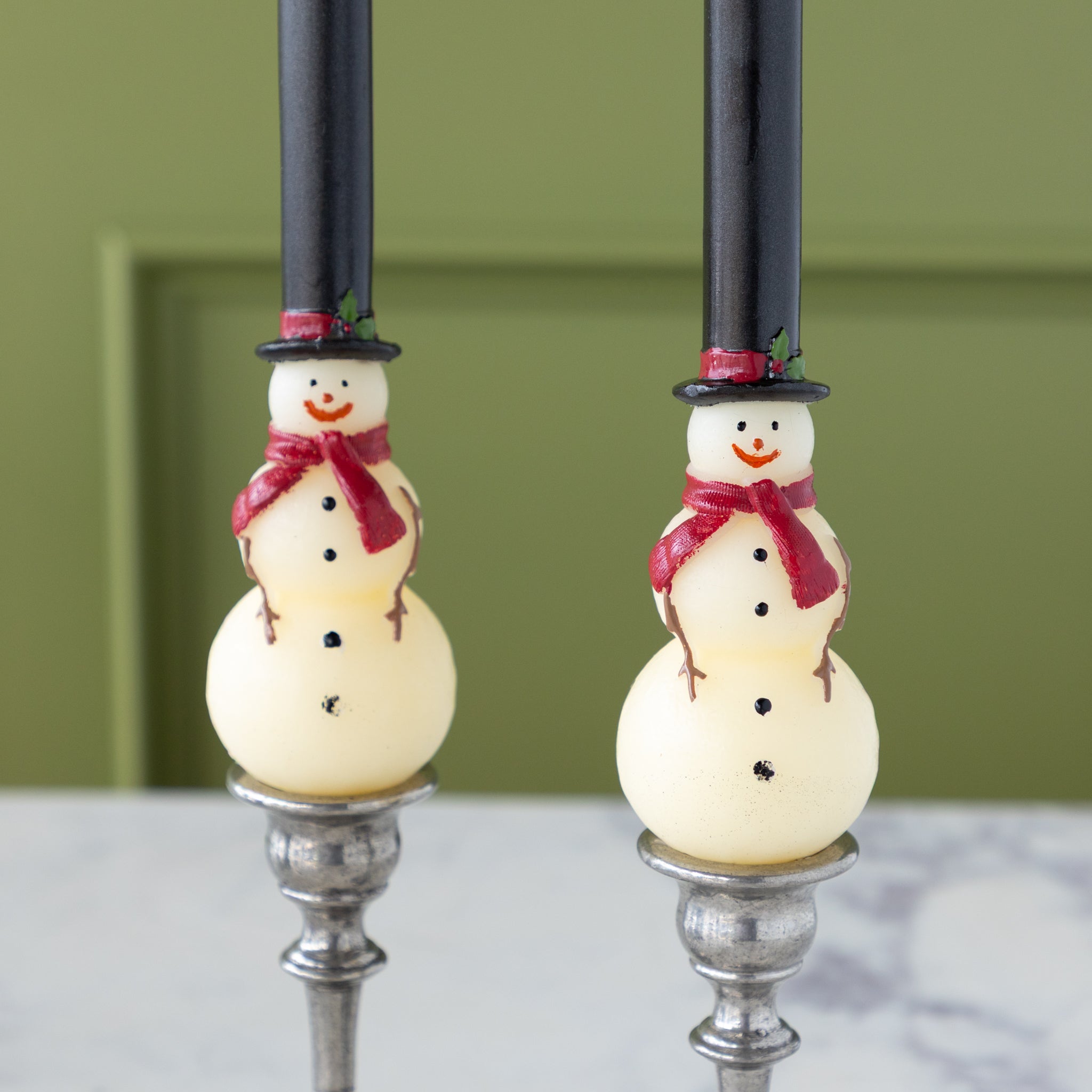 Two Snowman Taper Candles with tall black hats adorned with a red band and holly, red scarves, black buttons and smiley faces.