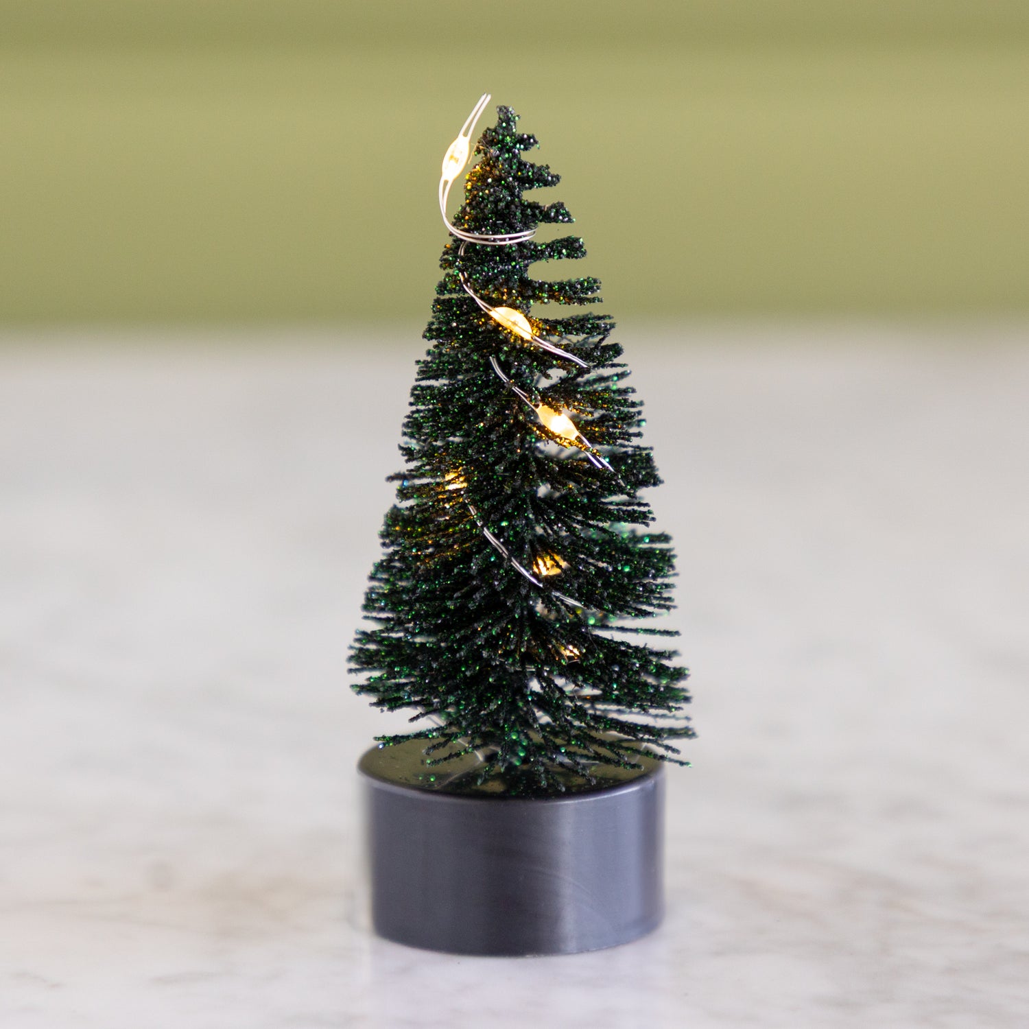 Small forest green bottle-brush tree with lights on a neutral background.
