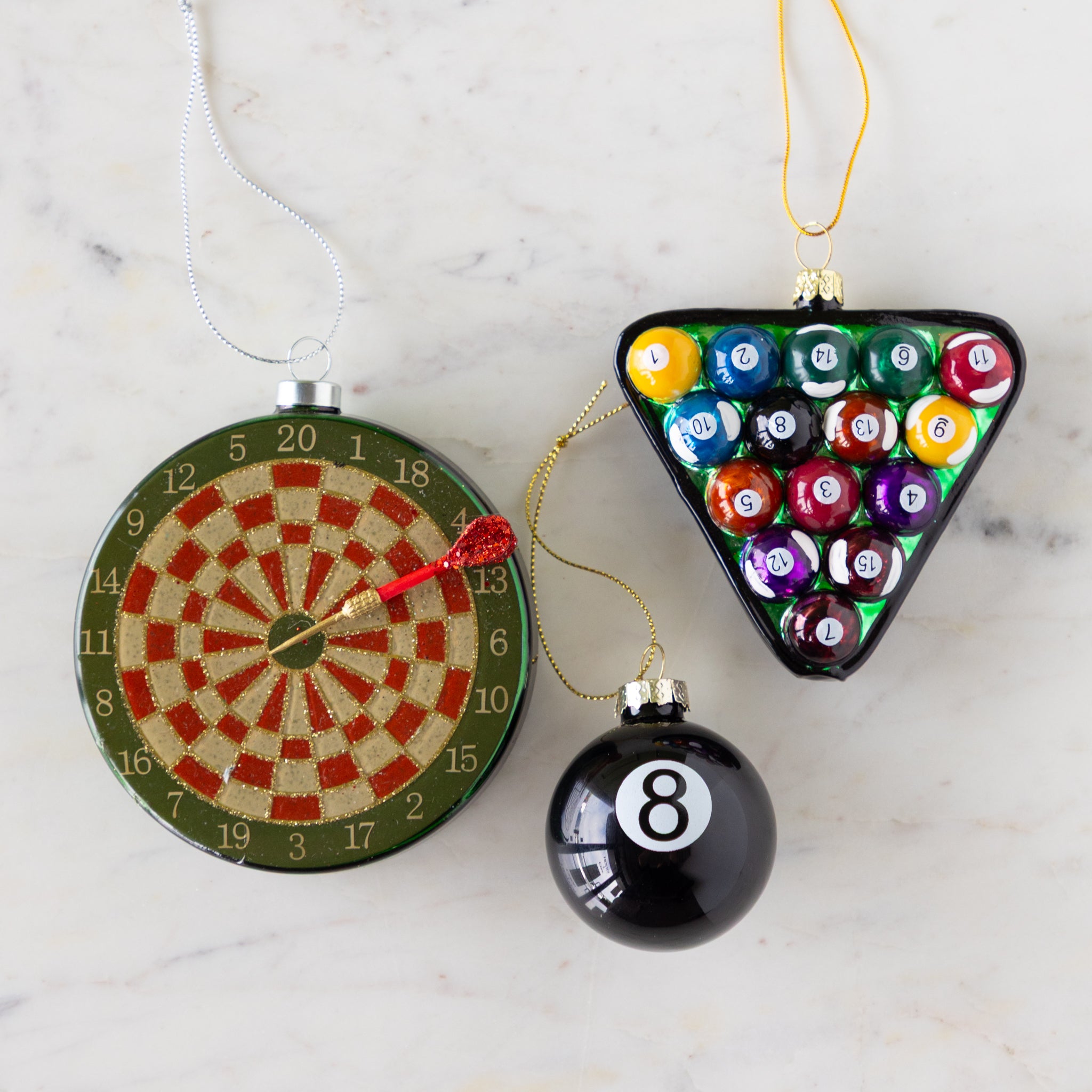 Three festive ornaments shaped like a dartboard, pool ball triangle, and number 8 ball on a marble surface.