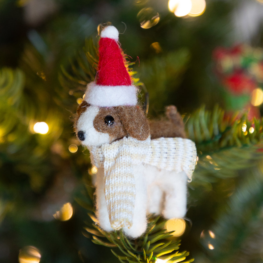 A small felt dog ornament wearing a red and white Santa hat and a white knit scarf, hanging on a green Christmas tree with warm blurred lights in the background.