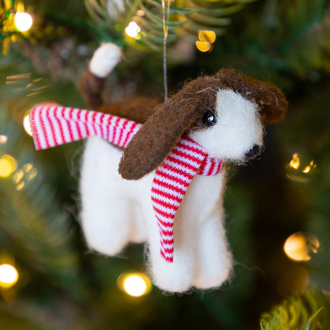 A small felt dog ornament wearing a red &amp; white  striped knit scarf, hanging on a green Christmas tree with warm blurred lights in the background.