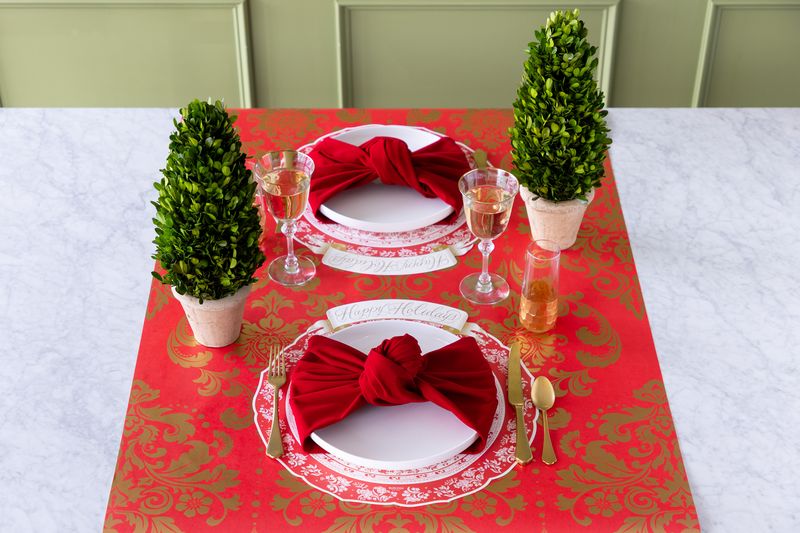 Table setting with red and gold paper table runner, white plates, red napkins, and green topiary plants.