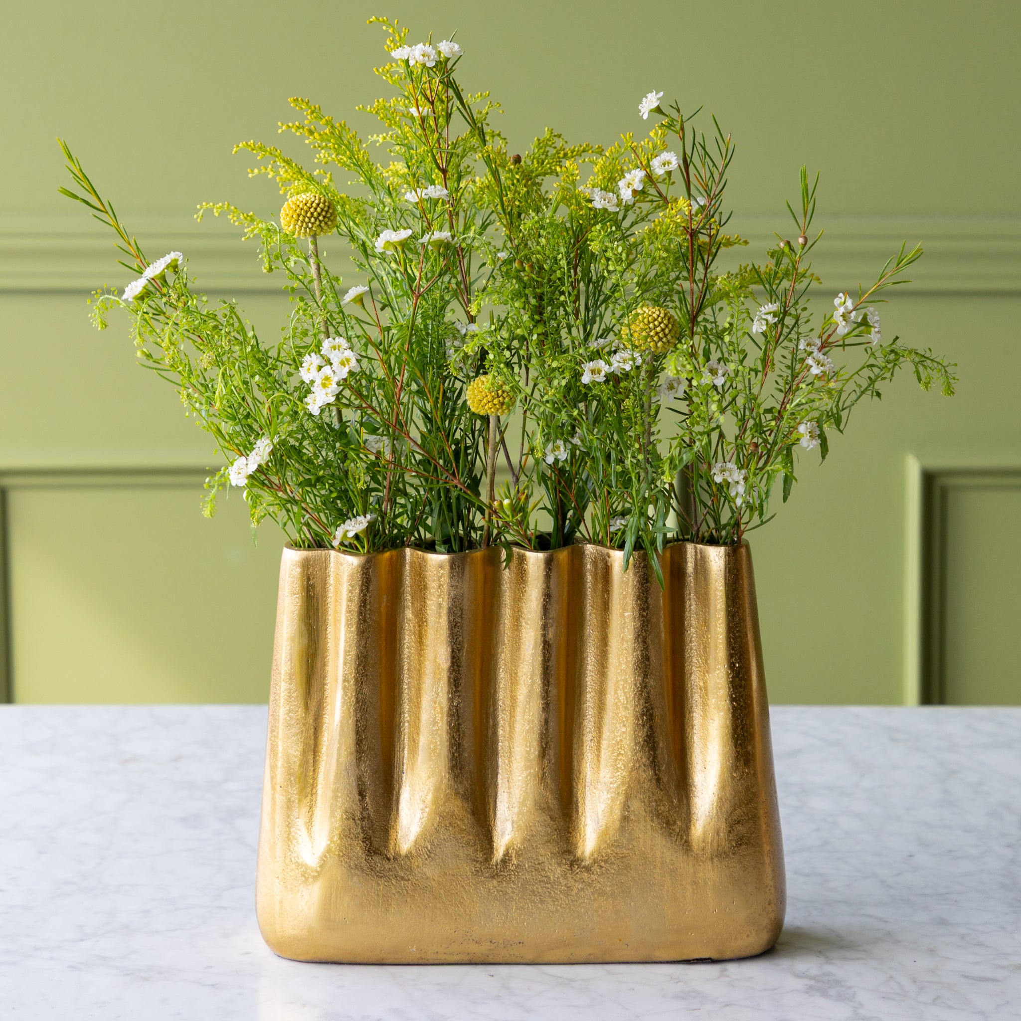 Tall Aluminum Gold Wave vase with greenery on a light surface against a green wall.