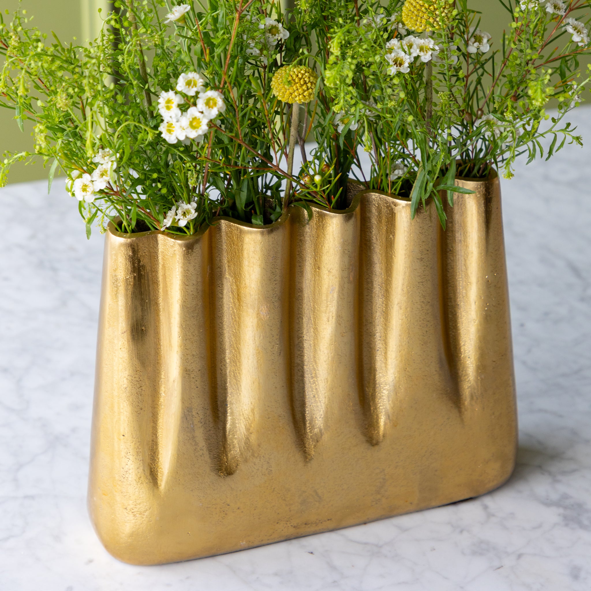 Tall Aluminum Gold Wave vase with greenery on a light surface against a green wall.