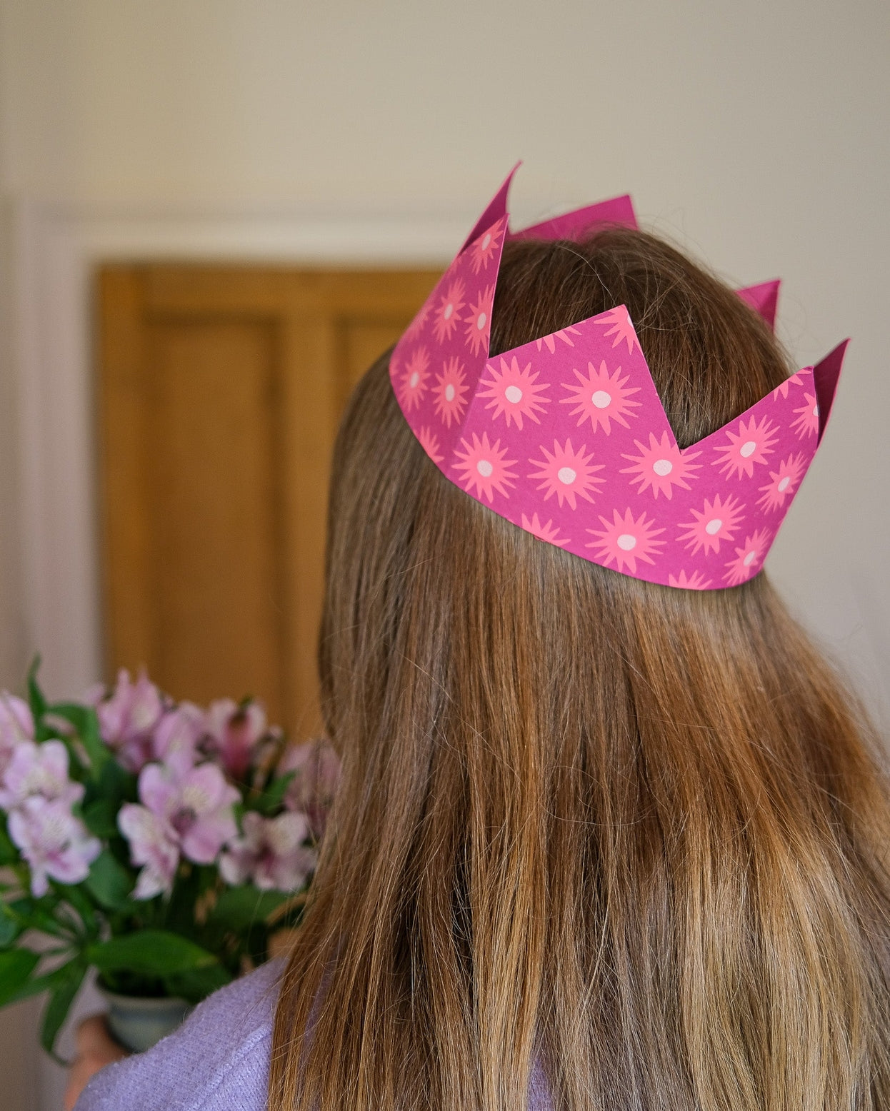 Person wearing a pink paper crown with floral patterns, standing indoors with a neutral background.