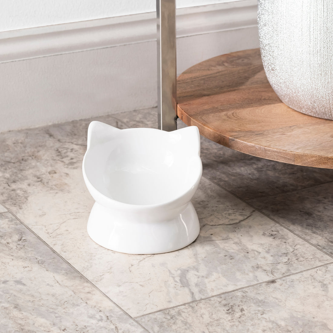 White cat-shaped food bowl on a marble floor with a wooden shelf in the background.