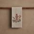 Tea towel with fox and mushroom design hanging on a wooden rod against a beige wall.