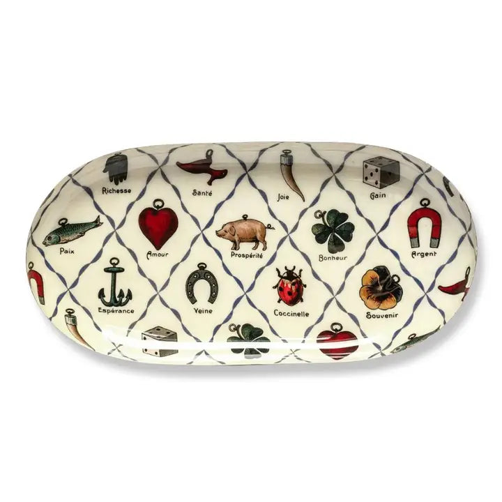 An oval shaped enamel plate containing a pattern of illustrated charms including a horseshoe, a lady bug, a four-leafed clover, and other symbols of luck. 