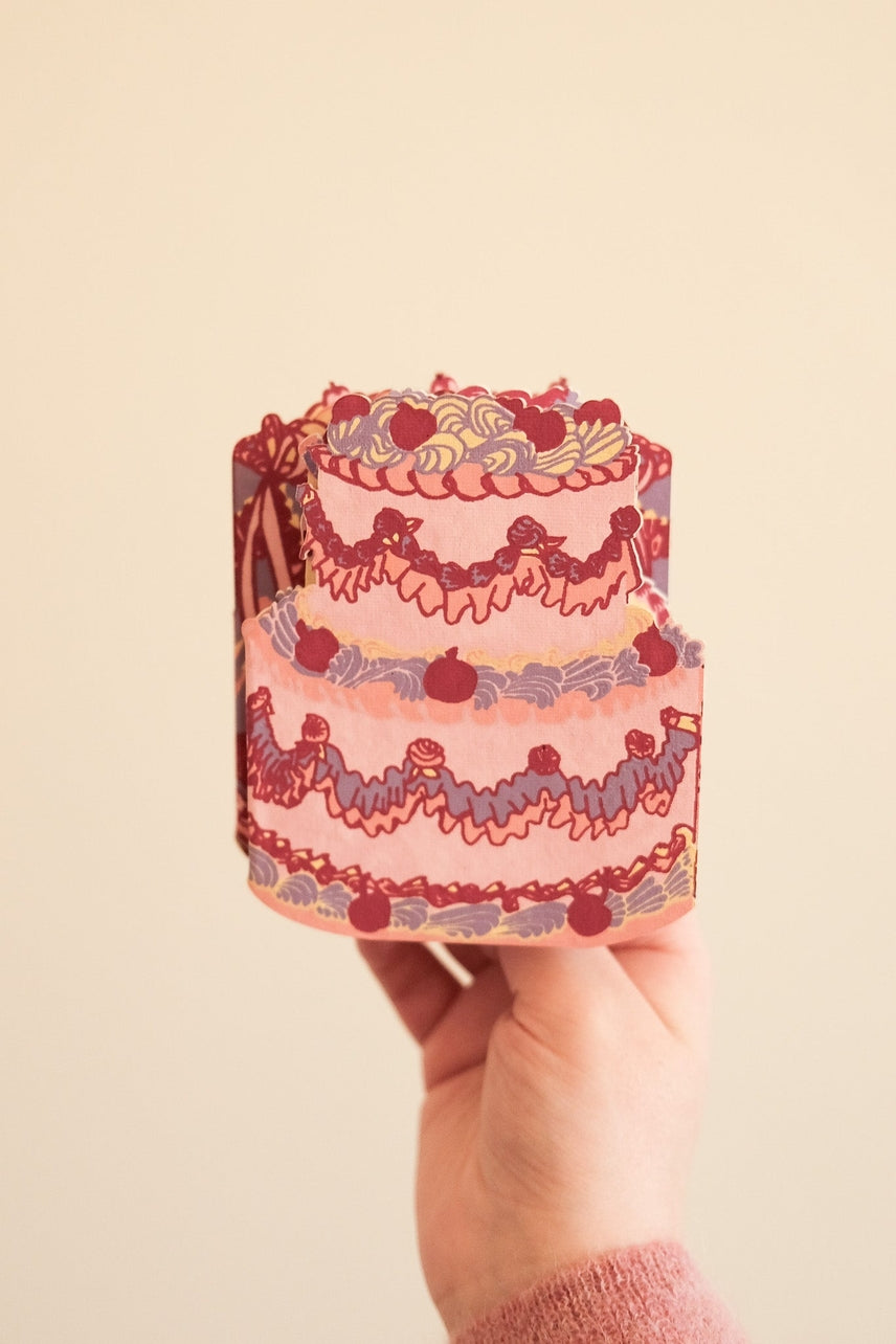 Hand holding a birthday cake shaped card against a beige background.