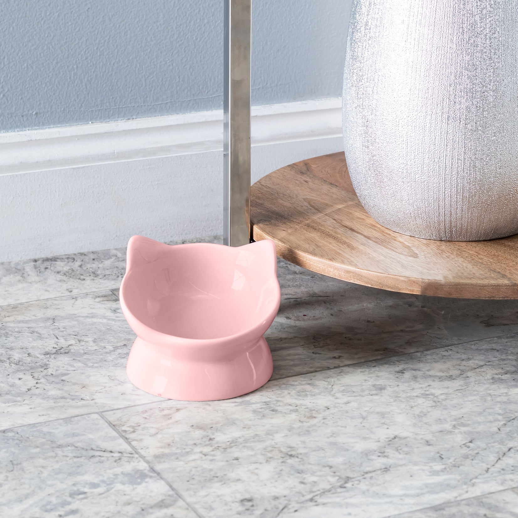 Pink cat-shaped bowl on a marble floor with a wooden shelf in the background.