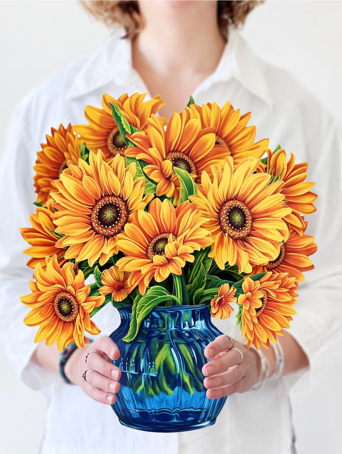 Person holding a bouquet of paper sunflowers in a blue vase against a white background. 