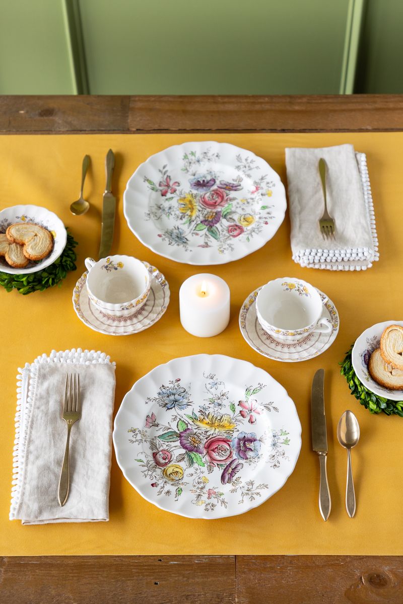 Set dining table with floral plates, cups, and cutlery on a golden yellow felted table runner.