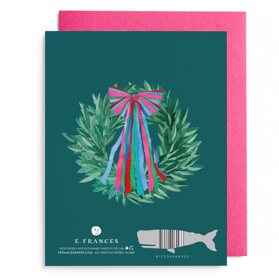 A square greeting card with a dark teal background featuring a watercolor-style green wreath and bright pink, blue, and red ribbon bow. A bright pink envelope is partially visible behind the card.