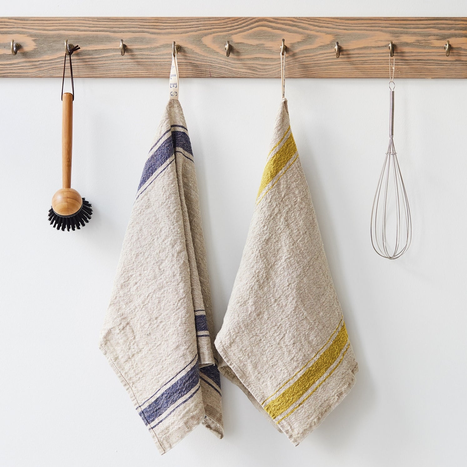 Two kitchen towels hanging on a wooden rack with a brush and whisk.
