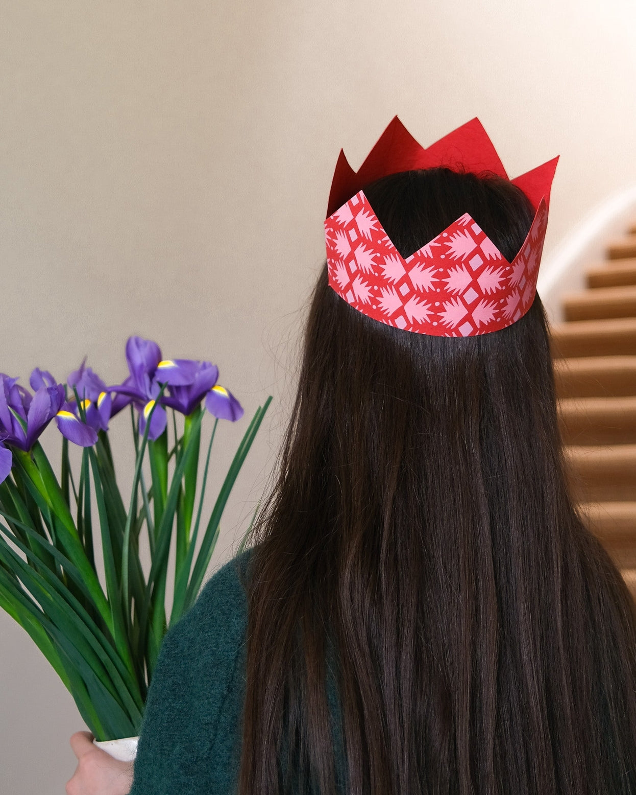 Person wearing a red paper crown with white patterns, holding purple flowers against a neutral background.