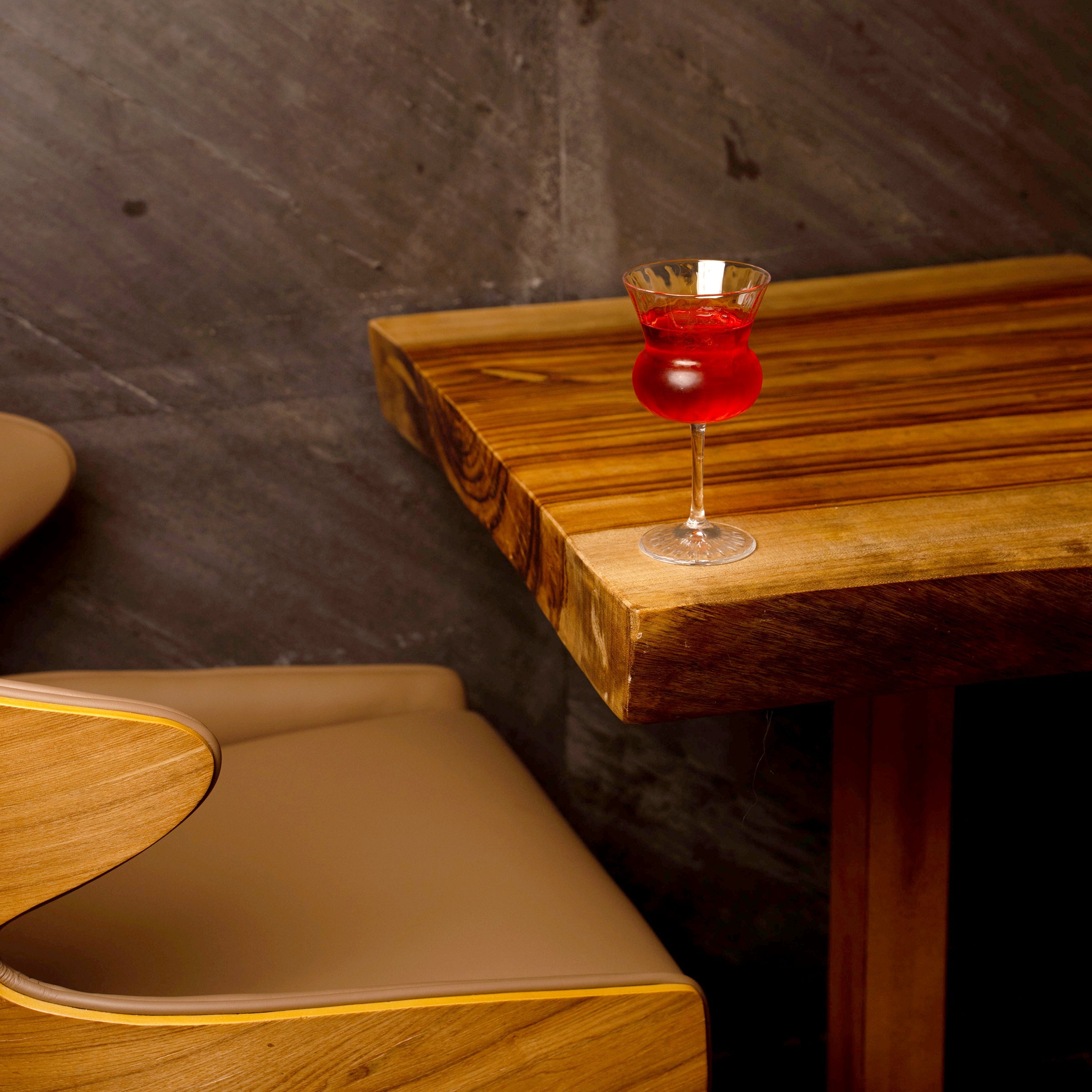 A stemmed glass filled with a bright red drink sits on a rustic wooden table in a dimly lit restaurant or bar setting with a dark textured wall and tan upholstered seating.