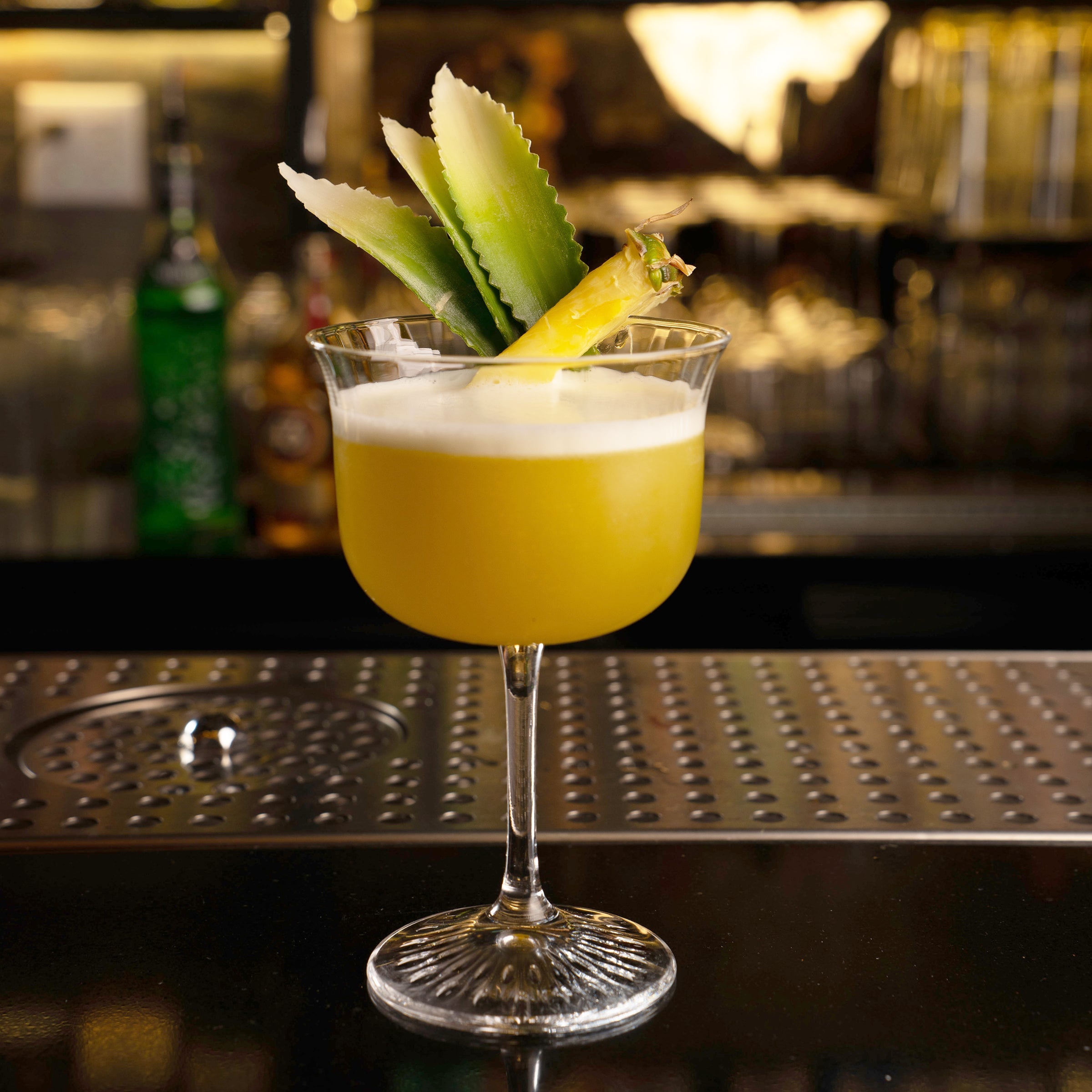 A yellow cocktail in a coupe glass with a white foam head, garnished with pineapple leaves and a pineapple core, set on a bar counter with a blurred bar background.