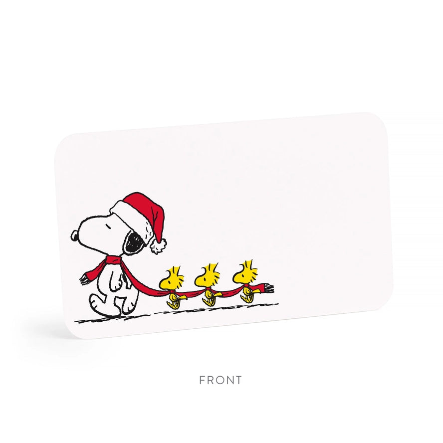 A white rectangular card or label with rounded corners, shown at a slight angle on a white background. The card features a printed illustration of Snoopy wearing a red Santa hat and scarf, pulling three yellow Woodstock figures.