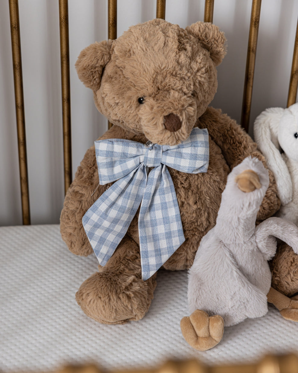 Brown teddy bear with a blue checkered bow tie sitting on a crib with a white blanket.