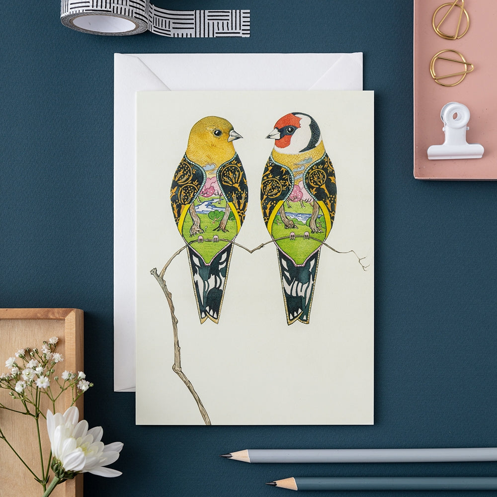 Greeting card with two stylized birds on a branch, placed on a dark blue surface with stationery items.