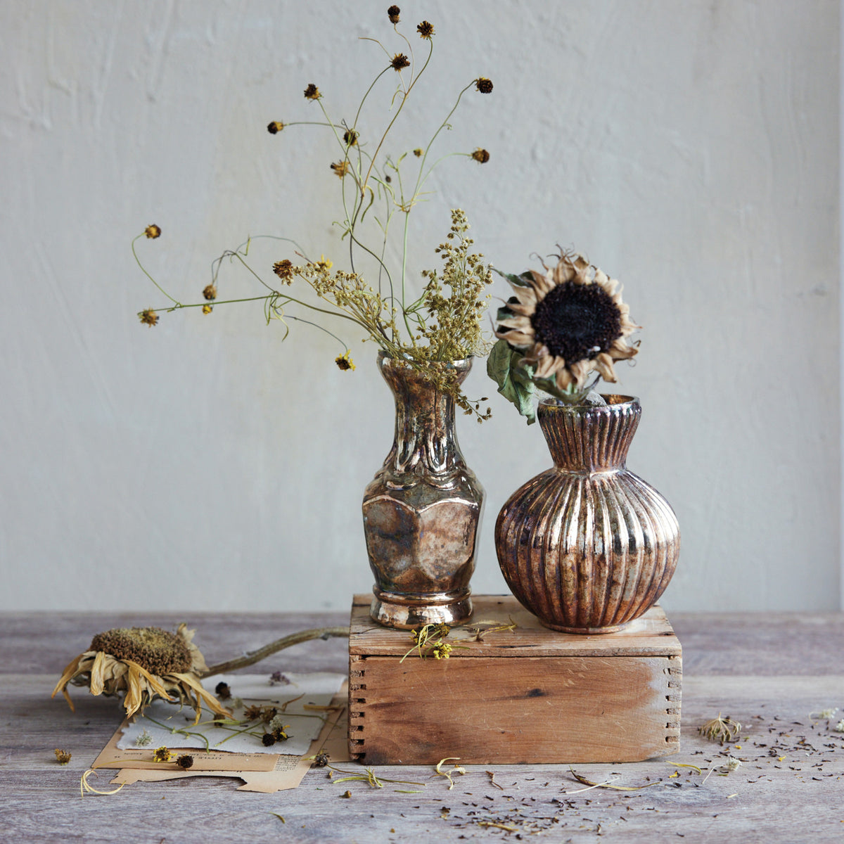 Two metallic vases with dried flowers on a wooden surface against a light gray wall.