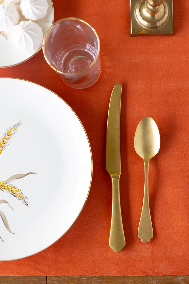 Table setting with white plate, gold cutlery, and napkin on an orange felted table runner.