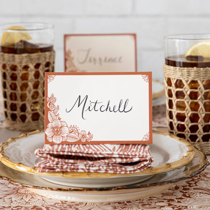 A free-standing, rectangular white table card with a dark orange frame around the edges, embellished with dark orange linework flower designs in the corners.