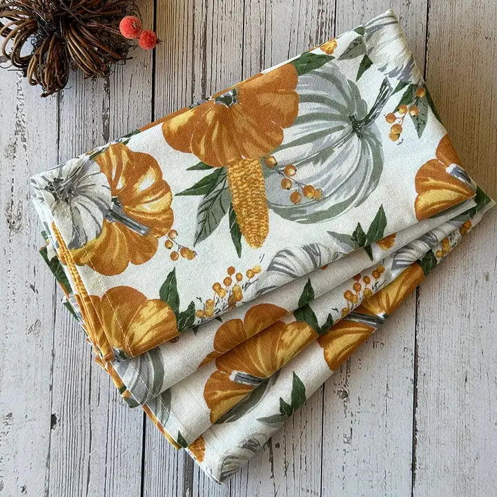 A folded fabric napkin featuring a fall harvest pumpkin and corn pattern on a cream background, styled on a rustic white wooden surface with a pinecone and small red berries.