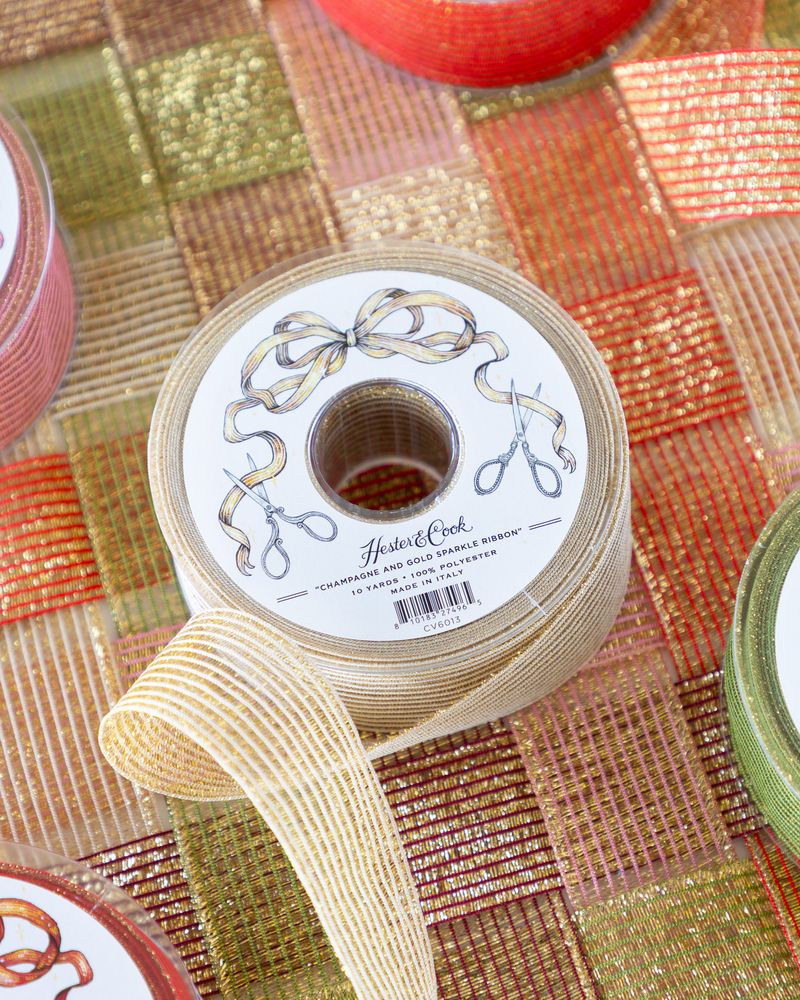 A spool of sparkly cream and gold ribbon on a surface of woven sparkle ribbon in five different colors. 
