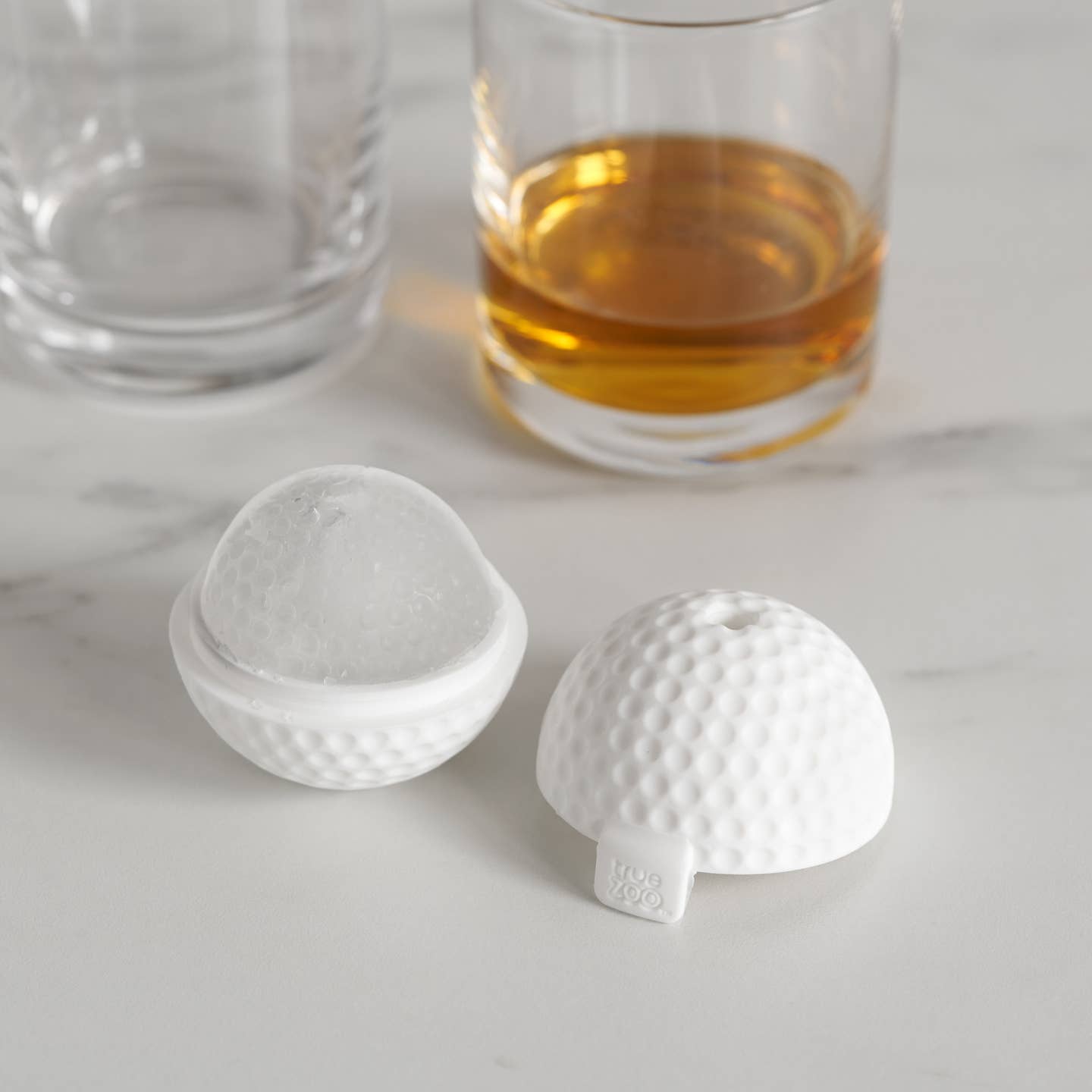 Two white silicone ice molds shaped like golf balls on a marble surface with glasses of liquid in the background.