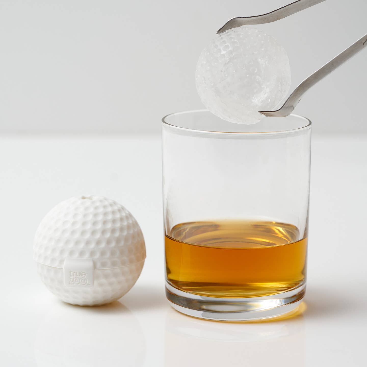 A golfball shaped ice cube next to a golfball shaped ice cube going into a glass of liquid.
