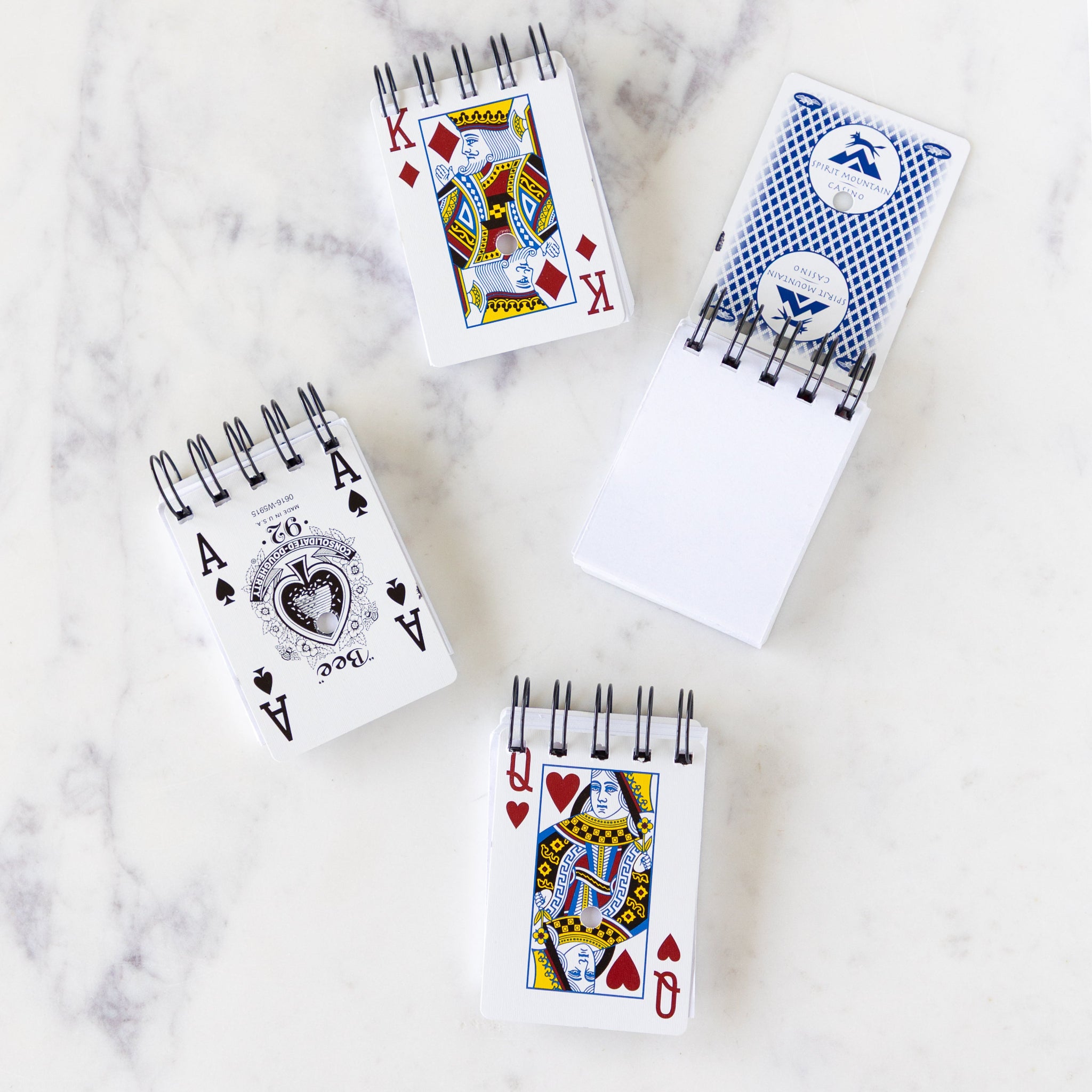Four Deck of Paper Notebooks on a marble table, each with a different paying card; one is open to show the blank page inside.