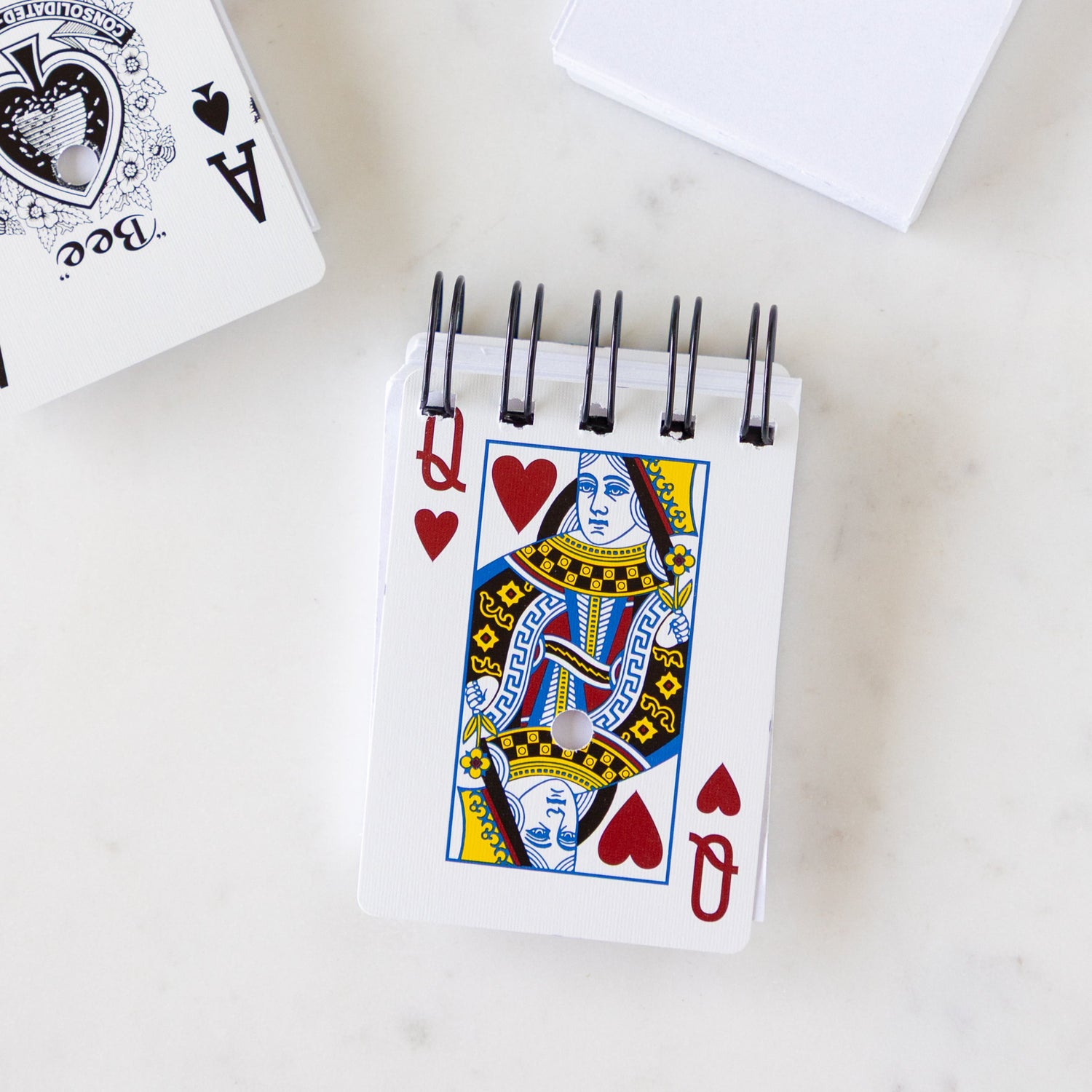 Small Deck of Paper Notebook with a Queen of Hearts playing card cover and a spiral bound, on a marble table.