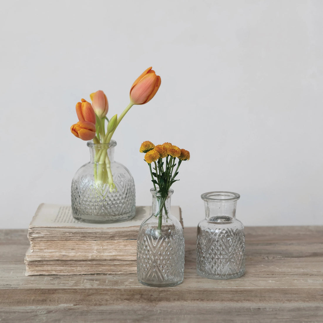 Three glass vases with flowers on a wooden surface against a light background.