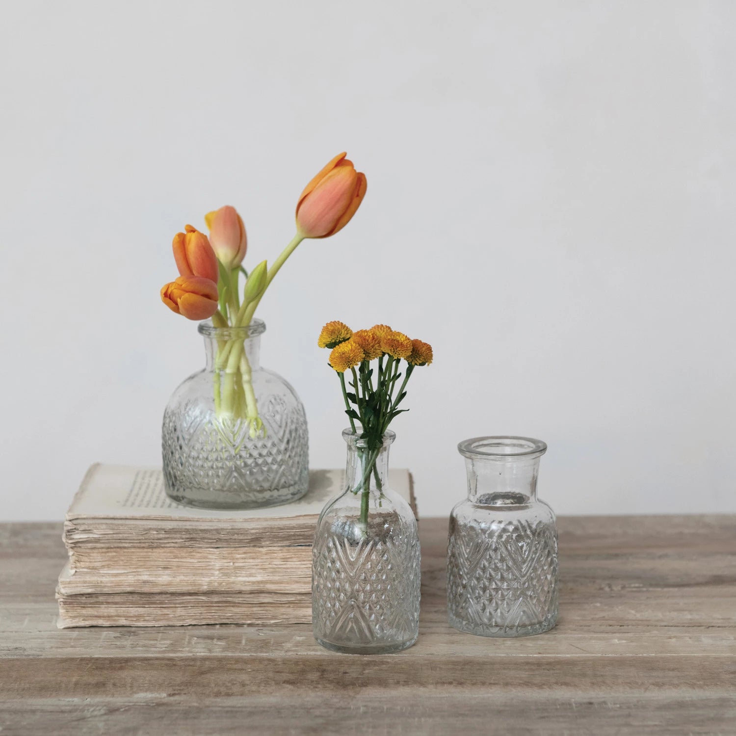 Three glass vases with flowers on a wooden surface against a light background.