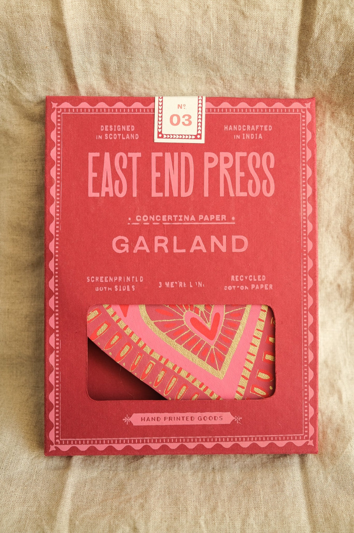 Red packaging of East End Press Garland product on a beige fabric background.