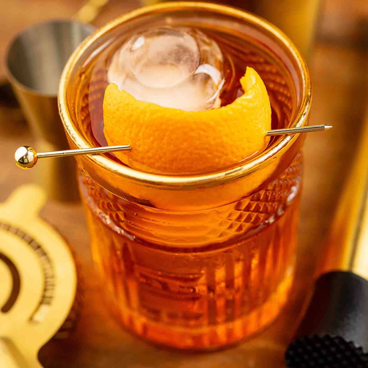 Close-up of a cocktail with a Gold Cocktail Pick piercing an orange peel garnish in a glass.
