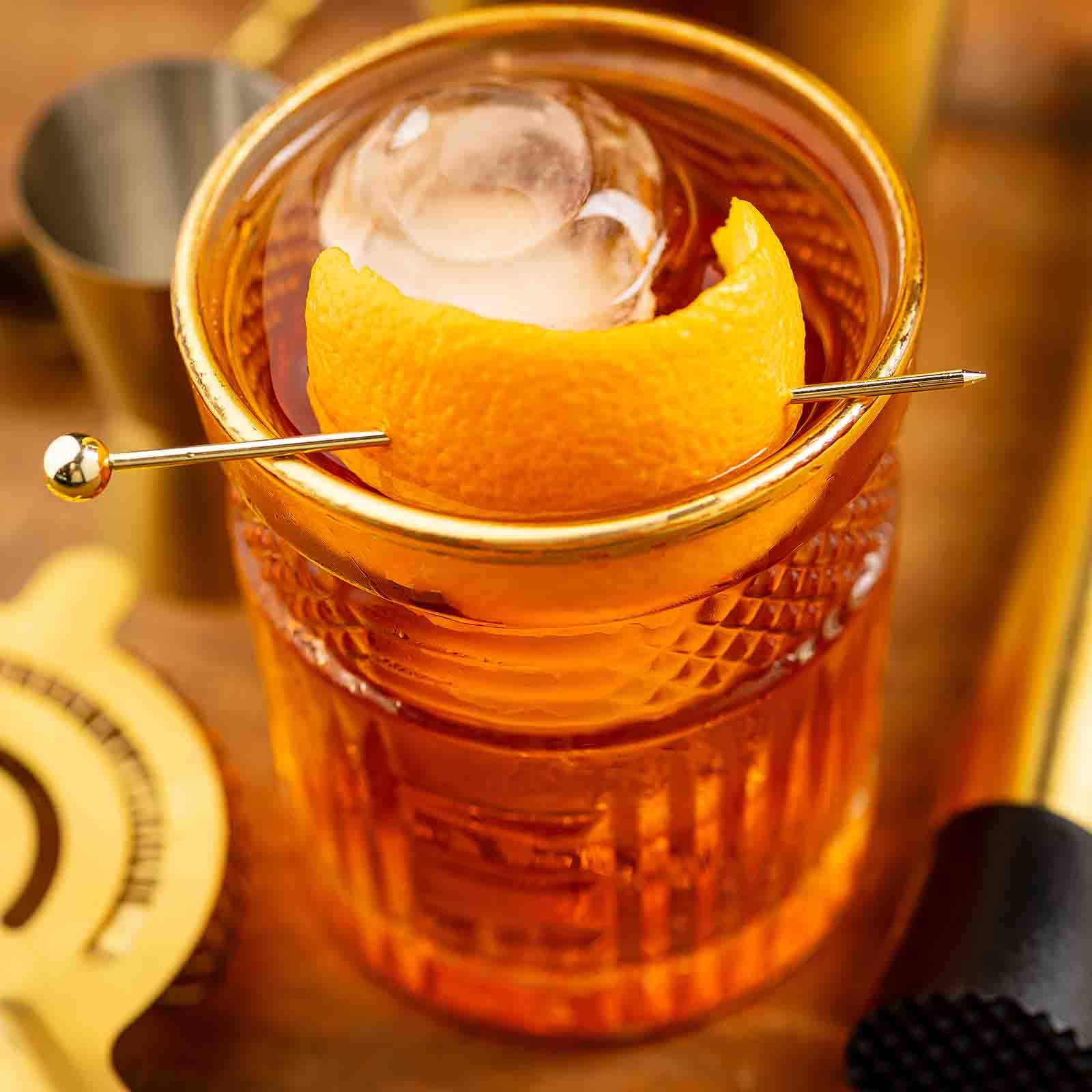 Close-up of a cocktail with a Gold Cocktail Pick piercing an orange peel garnish in a glass.