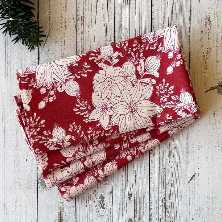 A folded red fabric napkin with a white floral and botanical print, displayed on a rustic white-washed wooden surface with a small green pine branch in the corner.